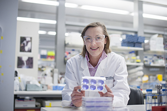 Professor Gabrielle Belz in laboratory holding experimental equipment, smiling at camera