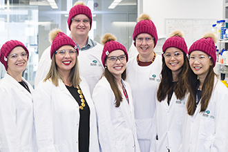 Large group of researchers in lab coats and beanies, standing and smiling at camera