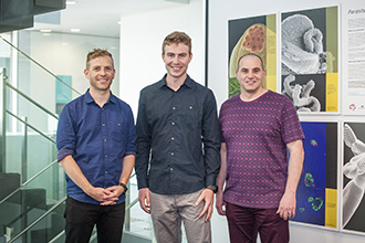 Three malaria researchers standing in front of brightly coloured posters, smiling