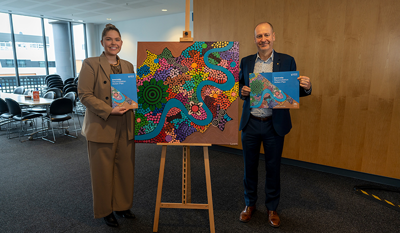 At the 2023 launch of WEHI’s third Reconciliation Action Plan, with First Nations artist Aimee McCartney (L). Artwork painted by Aimee McCartney is displayed in between the two individuals.