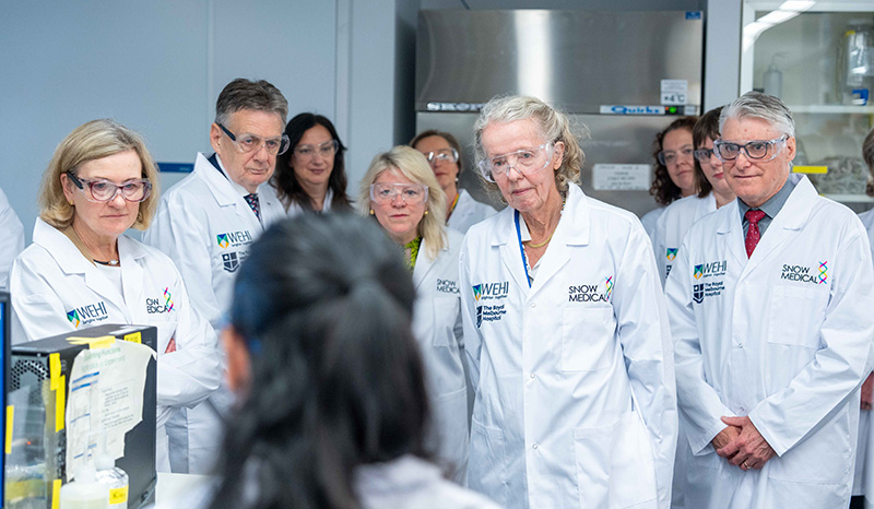A large group of people is photographed in a laboratory listening to a young researcher speaking