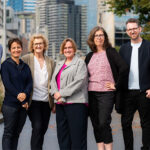 A group of 7 people photographed standing side by side in a park