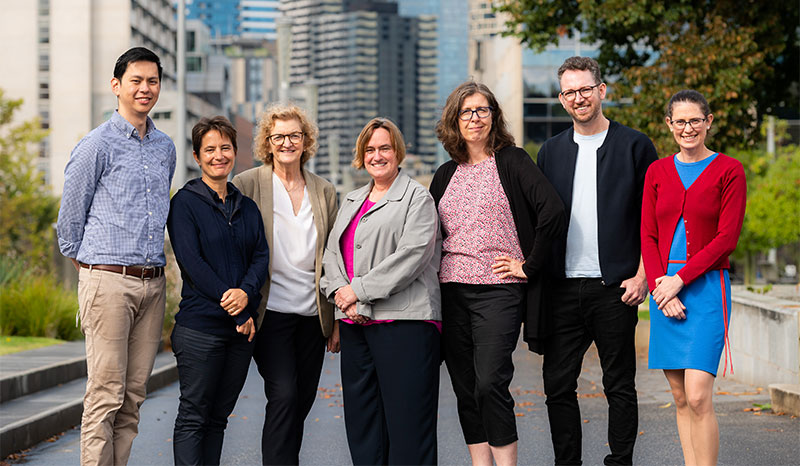 A group of 7 people photographed standing side by side in a park