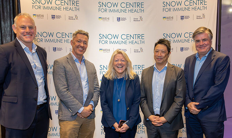 A group of five people are shown standing in front of a banner with the Snow Centre for Immune Health logo